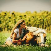 Young girl sitting on the grass caressing her lying horse